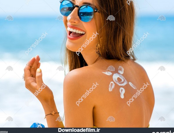 زن جوانی با شکل آفتاب روی شانه که بطری کرم ضد آفتاب را در ساحل نگه ...