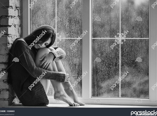 زن جوان زیبا تنها با قطرات باران نزدیک پنجره نشسته است و ...