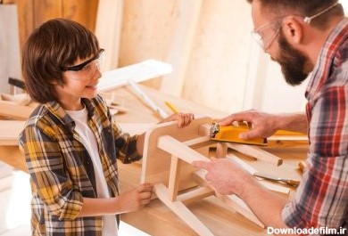 دانلود این به نظر می رسد شگفت انگیز است. پسر کوچک شاد به صندلی چوبی پدر خود در کارگاه کمک می کند