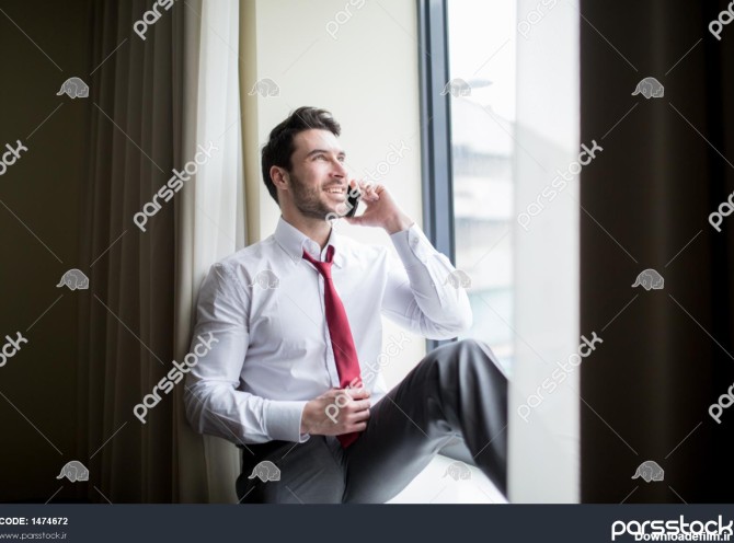مردی با لباس شخصی که در کنار پنجره نشسته و با تلفن همراه صحبت می ...