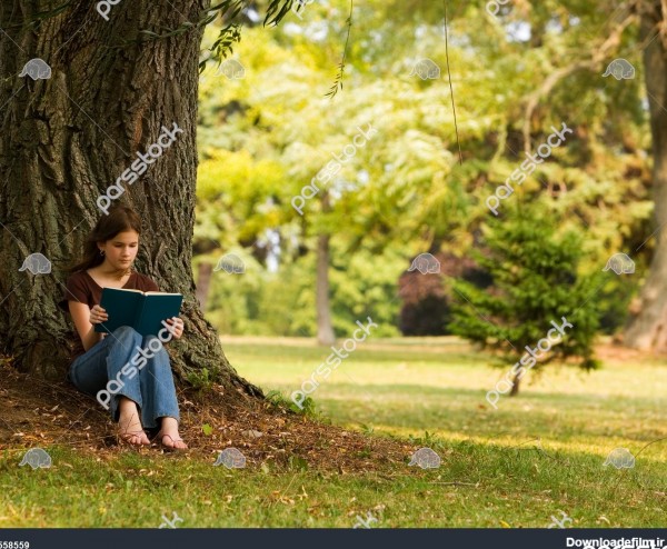 دختر دبیرستانی در حال خواندن کتاب در زیر درخت بزرگی در محیطی شبیه ...
