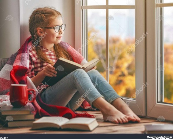 دختر بچه ناز نشسته در پنجره و خواندن یک کتاب در اتاق در منزل طبیعت ...