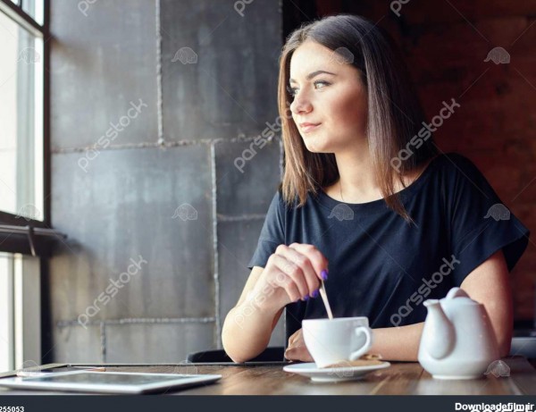 پرتره دختر جوان تکان دهنده قند در چای و متفکرانه از پنجره کافی شاپ ...